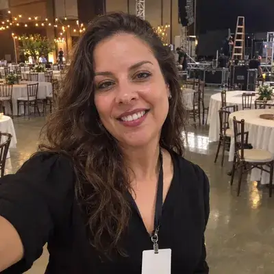 Selfie de Marcela, produtora de eventos, com crachá no pescoço em um salão de festas sendo montado, com mesas redondas e varal de luzes ao fundo