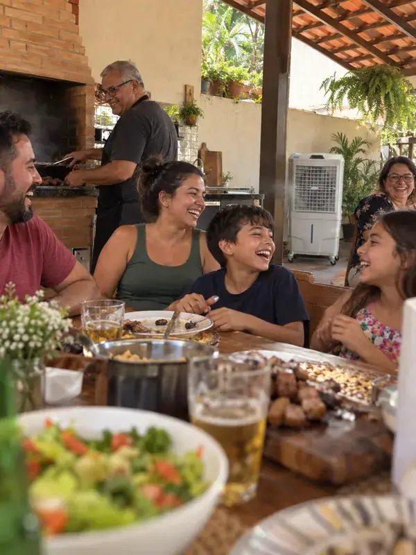 Família rindo à mesa de uma área gourmet enquanto o avô assa carne na churrasqueira, com um climatizador branco ligado ao fundo